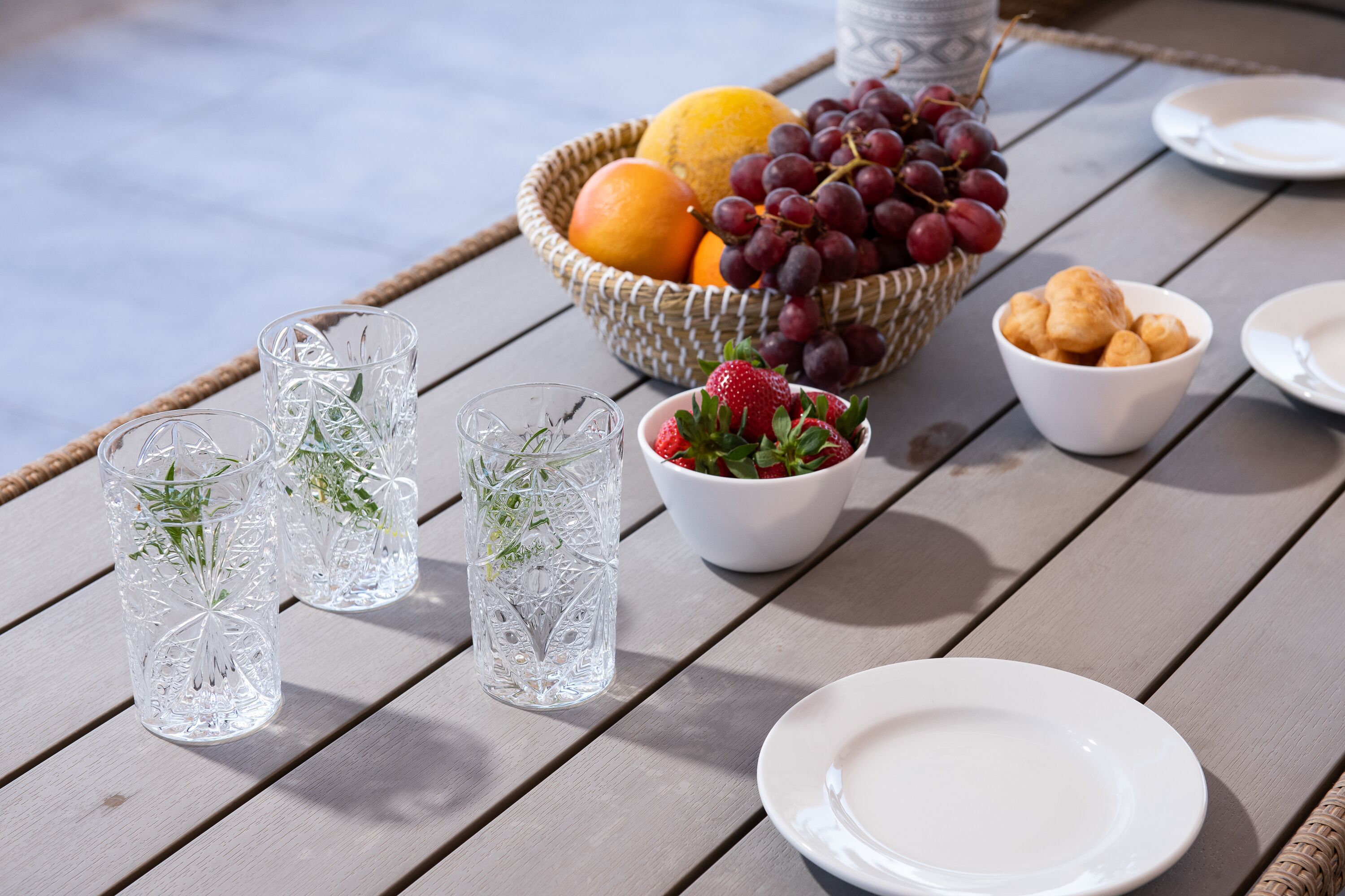 A table with a wicker basket containing fruits and grapes, a bowl of strawberries, and two clear glasses with herbs.