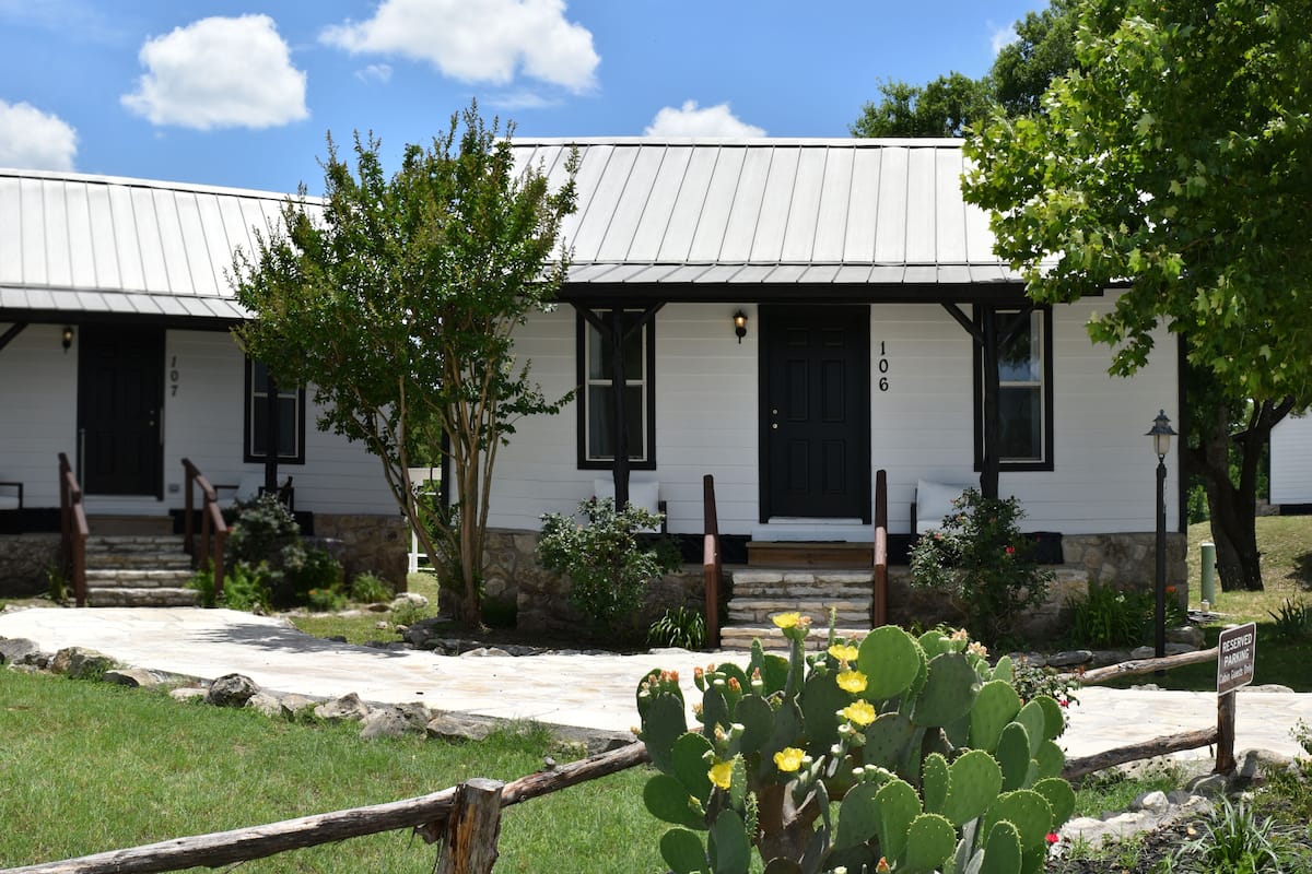 A white building with black-framed windows and doors, numbered 107 and 106, with a stone wall and steps leading to the entrance.