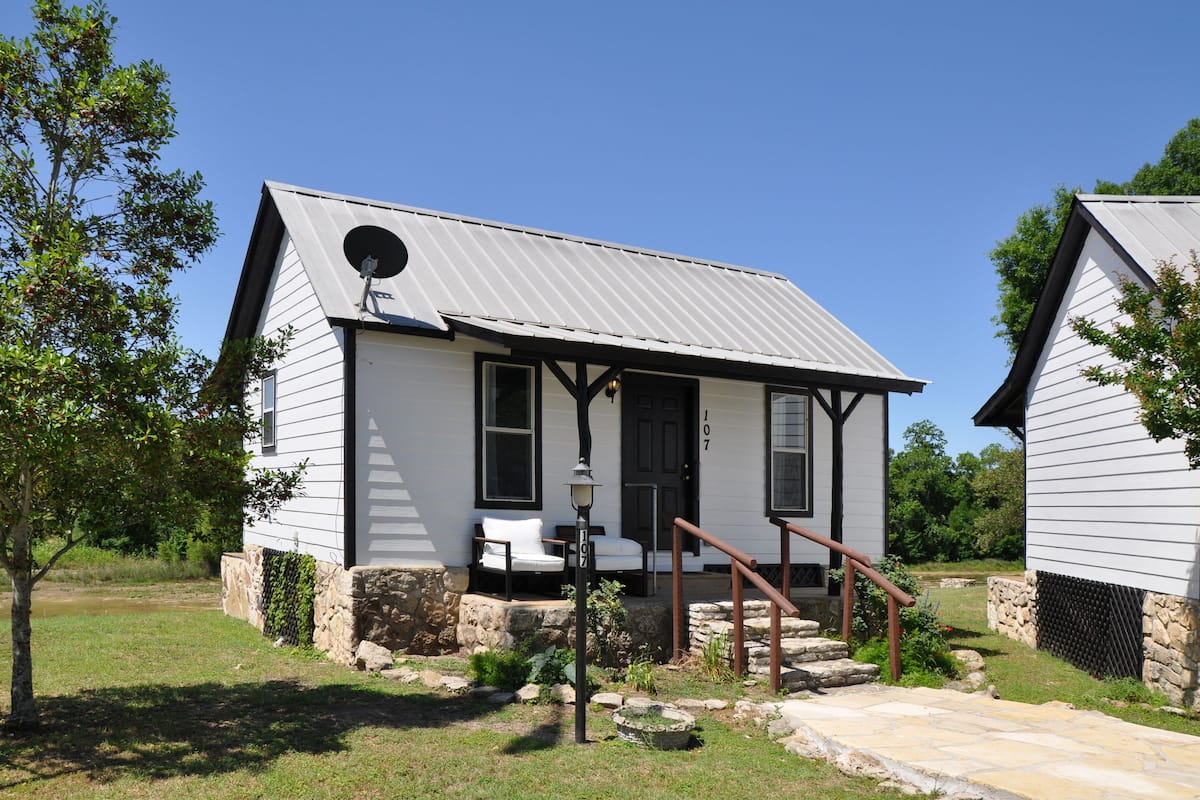 A white building with a metal roof, stone foundation, and a small porch with a bench.
