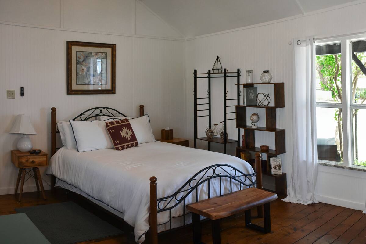 A bedroom with a wooden bed, a bench, a nightstand with a lamp, a shelf with decorative items, and a window with curtains.