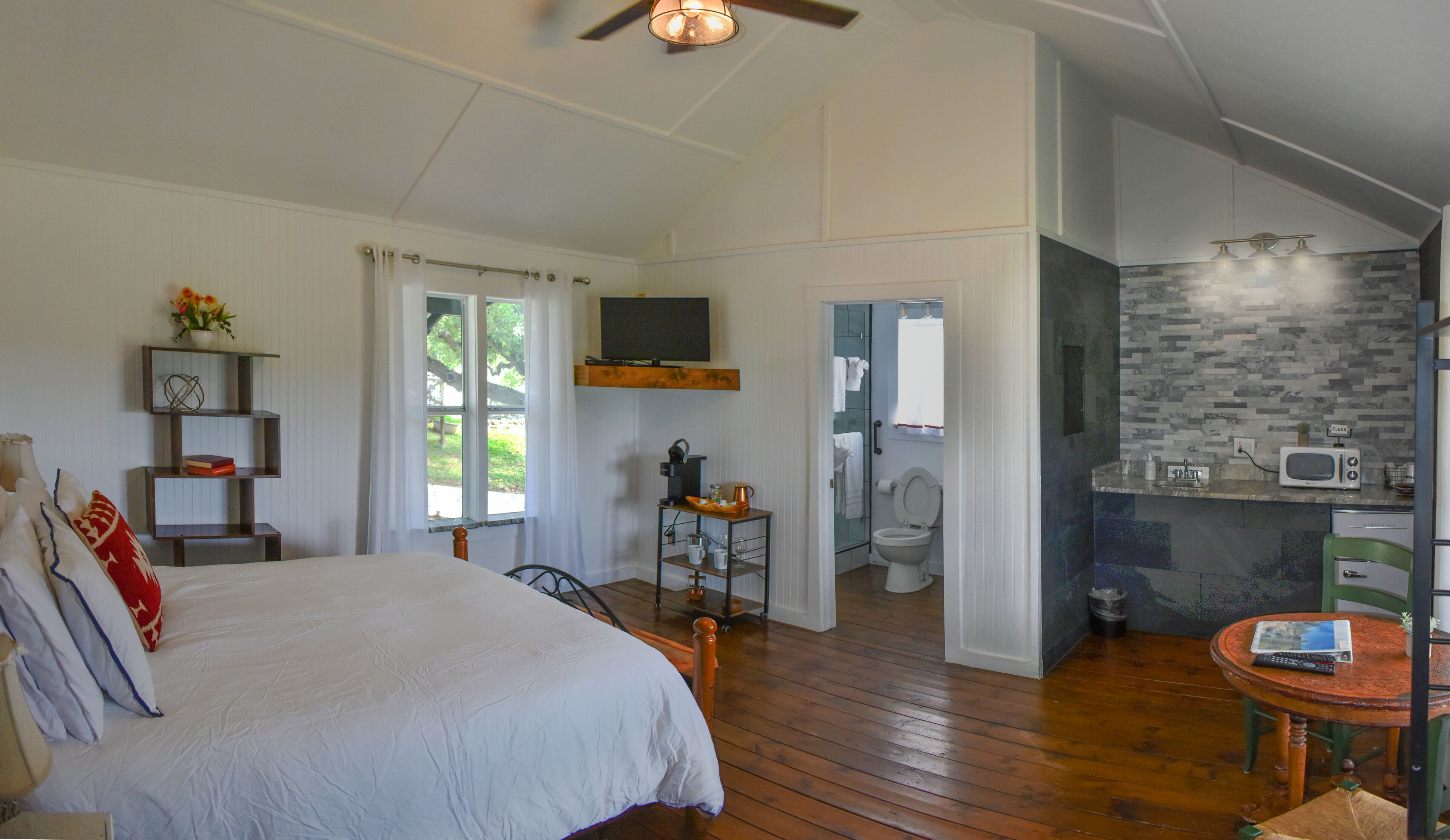 Ein Schlafzimmer mit einem großen Bett, einem Schreibtisch mit Stuhl, einer kleinen Küchenzeile mit Mikrowelle und einem Badezimmer mit Toilette.