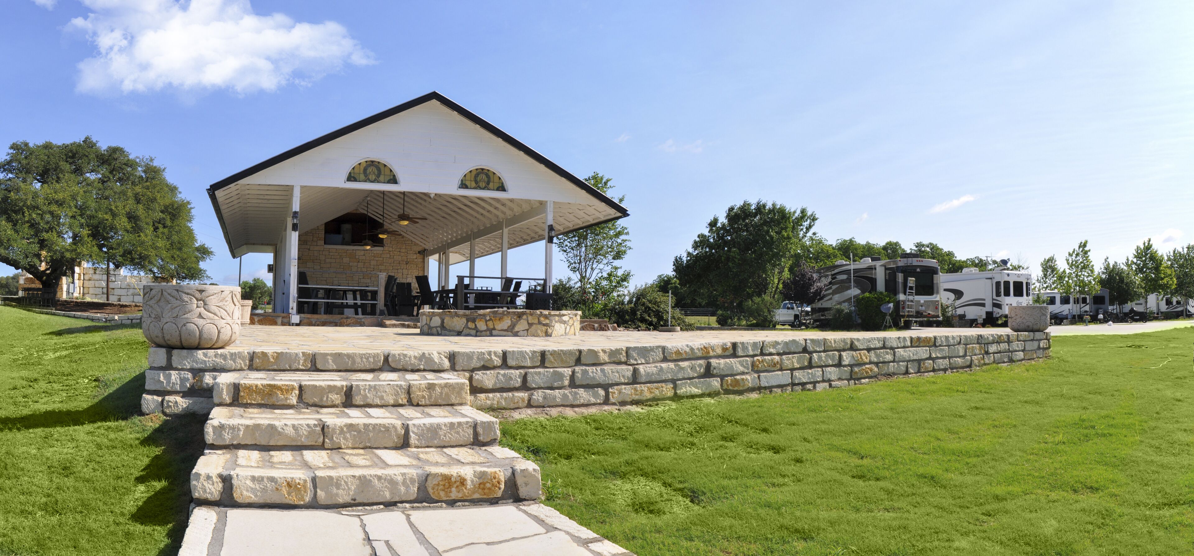 A covered pavilion with stone steps leading up to it, surrounded by a grassy lawn and trees.