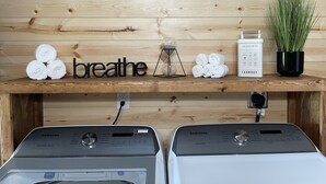 Et vaskerum med to Samsung vaskemaskiner, en hylde med håndklæder, et skilt med ordet 'breathe', en plante og en vejledning til vask.