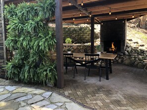 Un patio exterior con suelo de piedra, una mesa y sillas de madera, una pared de piedra y un brasero.