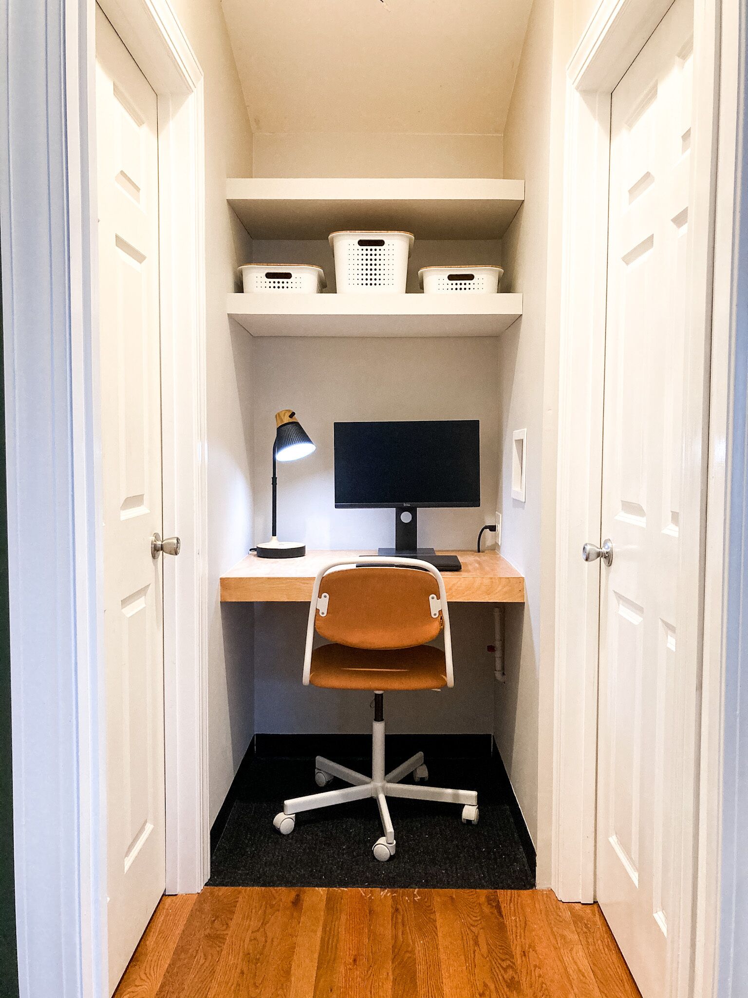 A small, narrow room with a desk, a computer, and a chair. There are shelves above the desk holding baskets.