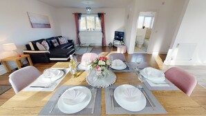 Spiseområde med et bord dækket til fire personer, en sofa, et fjernsyn og et vindue med gardiner.