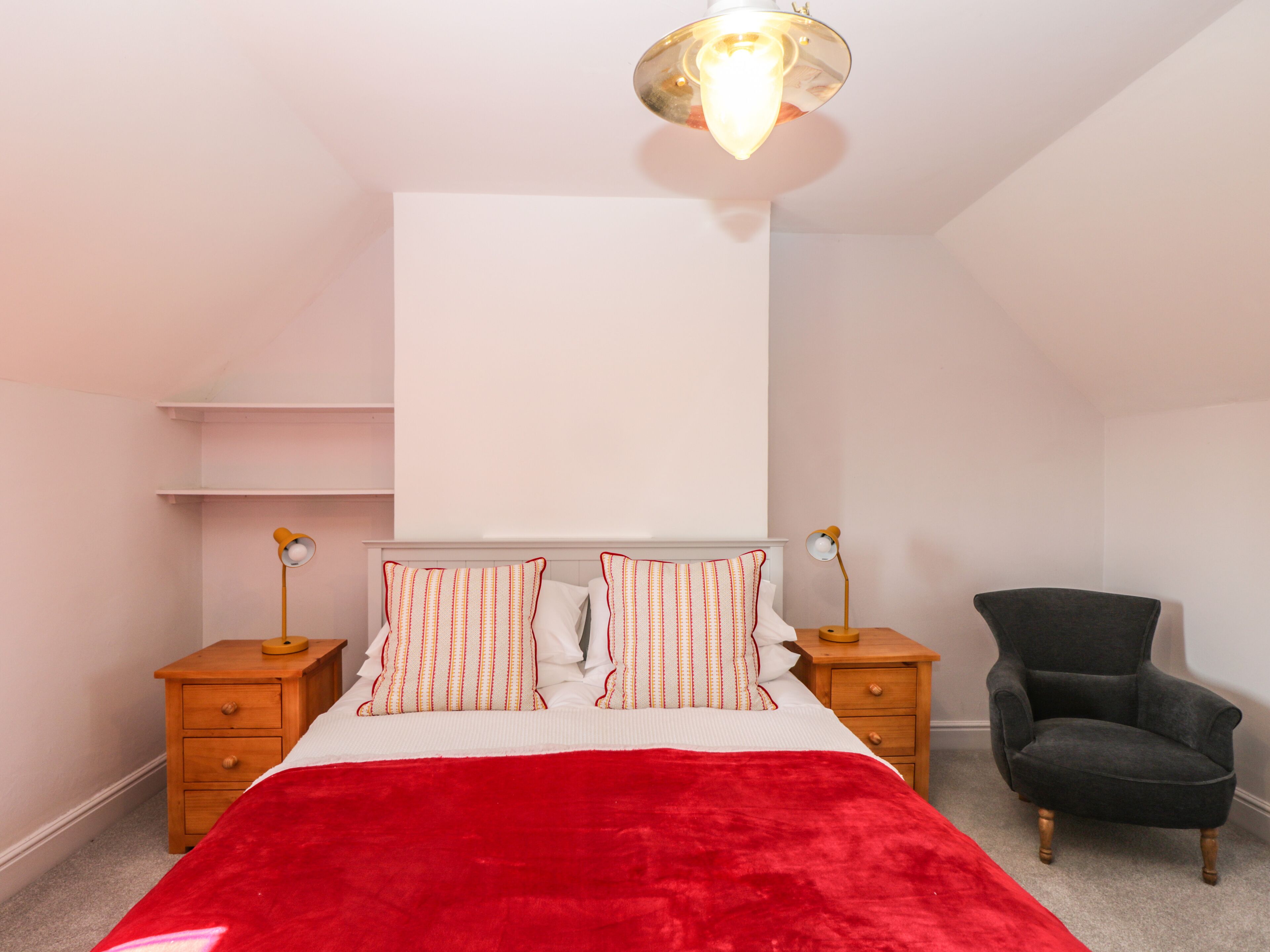 A bedroom with a bed, a red blanket, two bedside tables with lamps, a chair, and a white wall with a shelf.