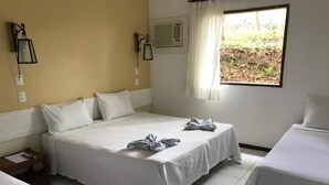 Une chambre d’hôtel avec un grand lit, deux petits lits, une fenêtre avec des rideaux et une petite table avec une lampe.