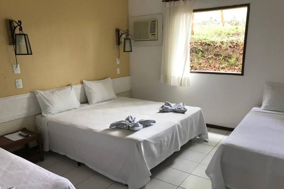 Ein Hotelzimmer mit einem großen Bett, zwei Einzelbetten, einem Fenster mit Vorhängen und einem kleinen Tisch mit einer Lampe.
