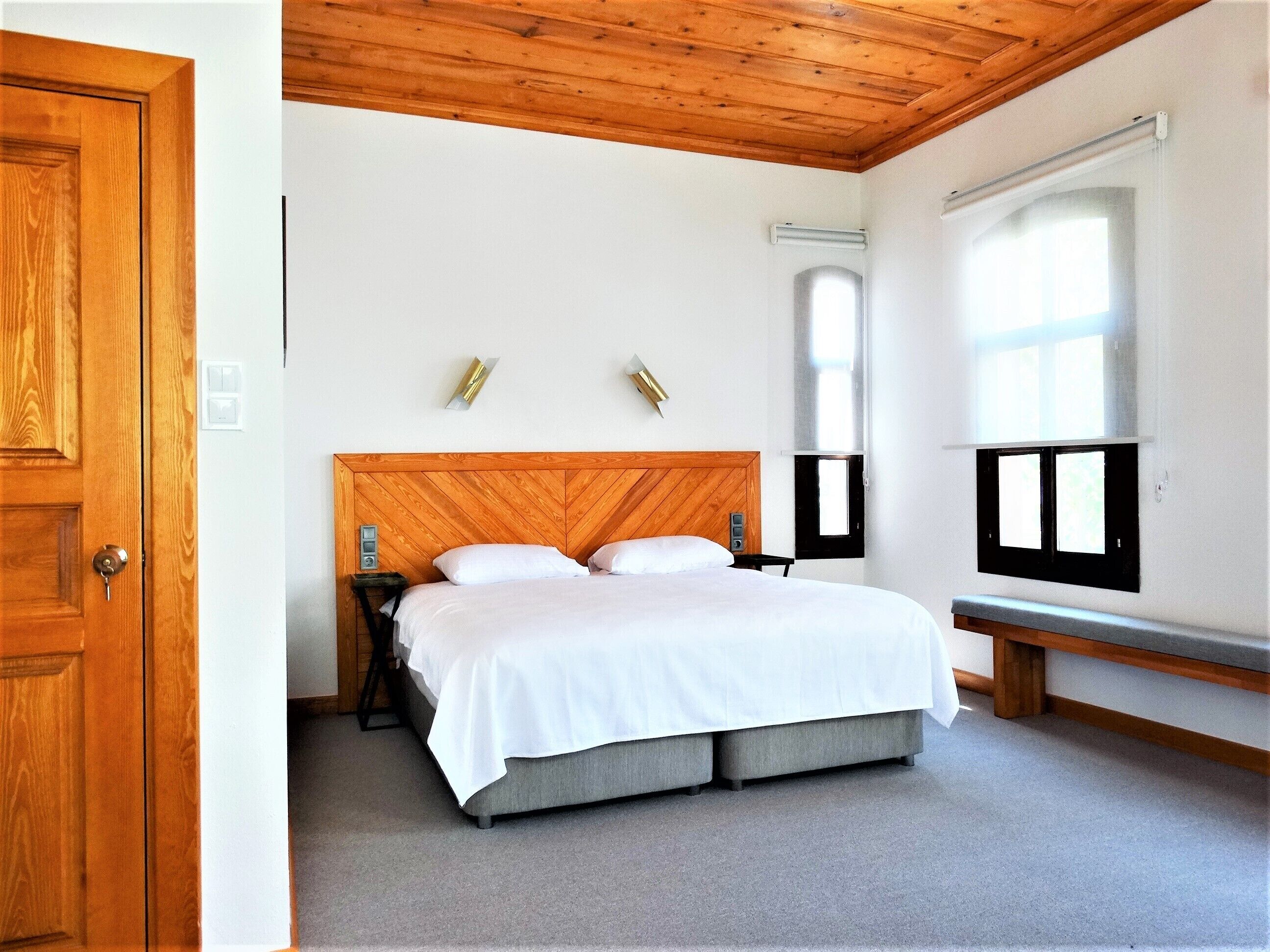 Une chambre avec une tête de lit en bois, un lit avec une literie blanche, une table de chevet, une commode, une fenêtre avec des stores et une porte.