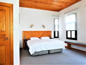 A bedroom with a wooden headboard, a bed with white bedding, a bedside table, a nightstand, a window with blinds, and a door.