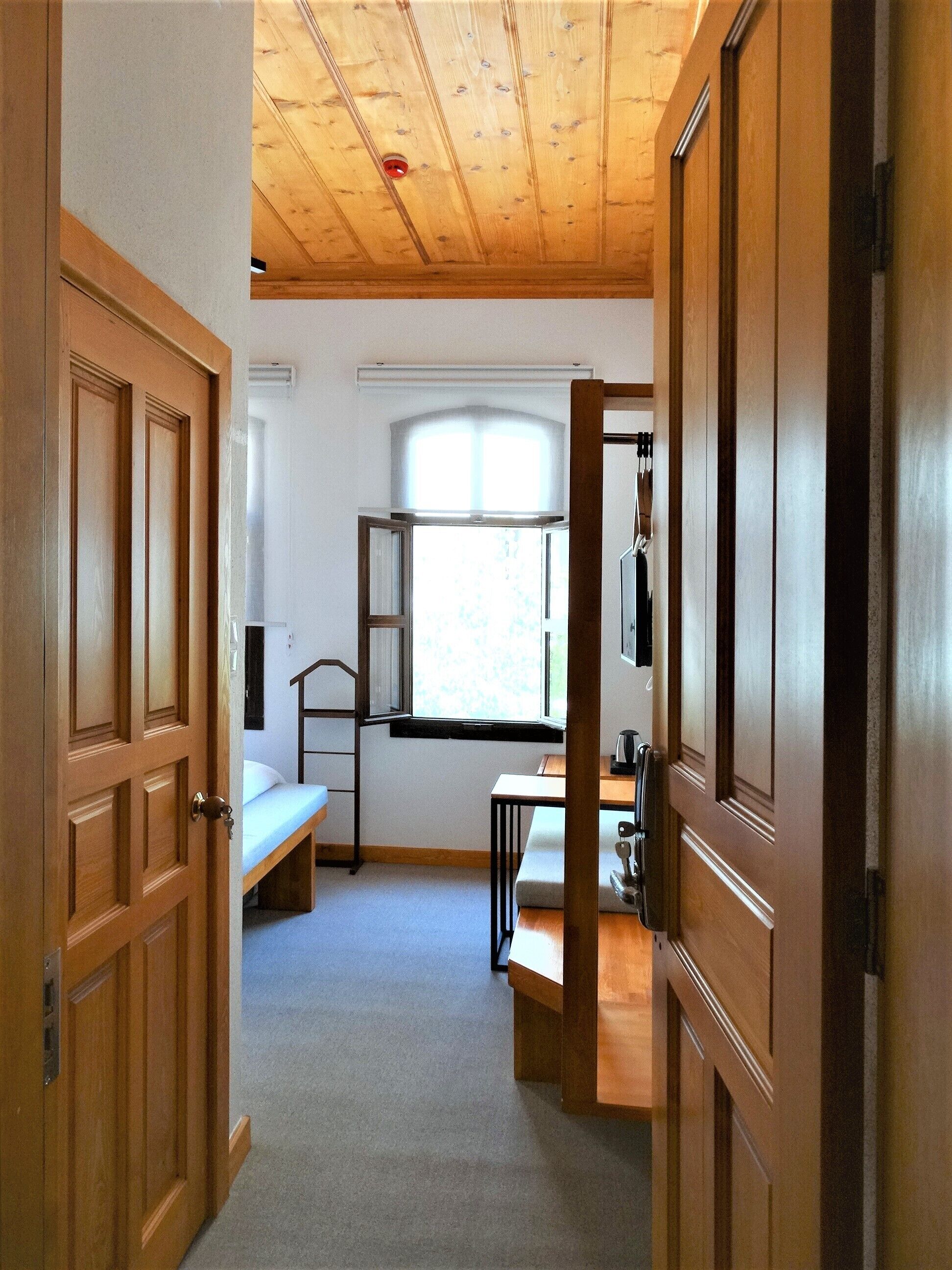 A room with a wooden ceiling, a window with a view, a bed, a desk, and a wardrobe.