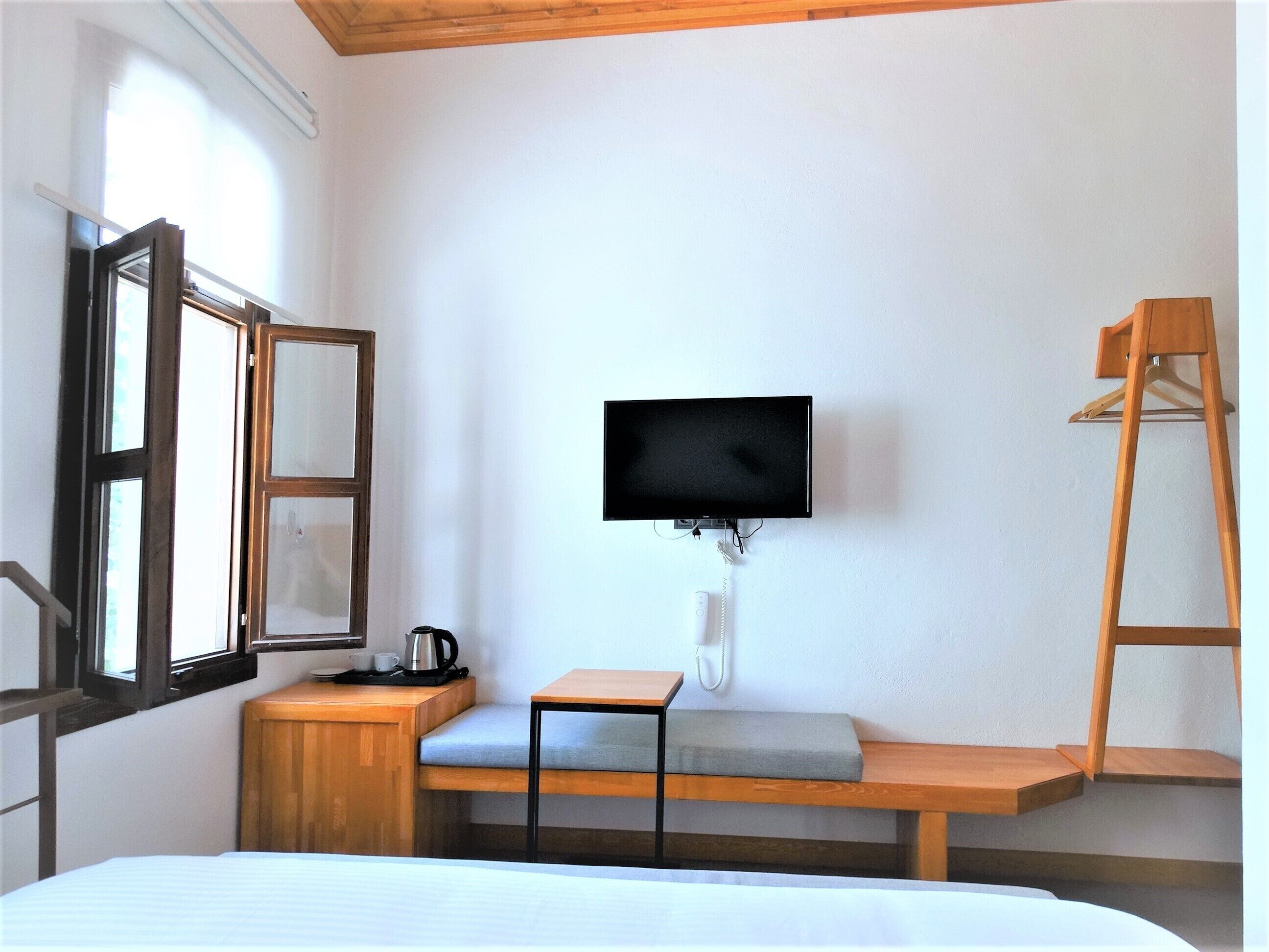 A room with a bed, a TV mounted on the wall, a wooden bench, a small table, and a window with a view of the outside.