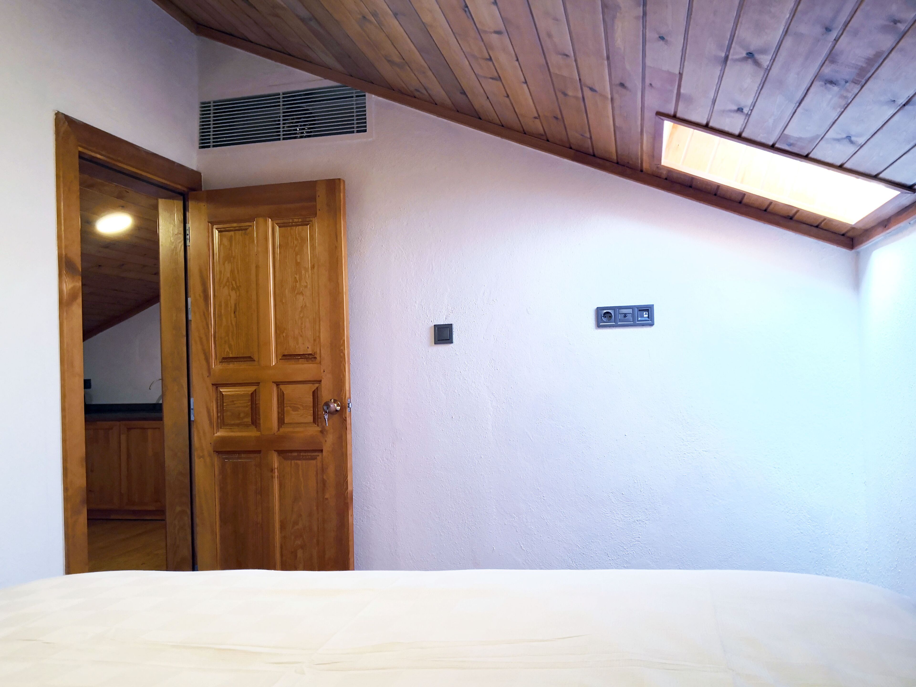 A bedroom with a wooden door, a bed, a ceiling vent, and a light switch on the wall.