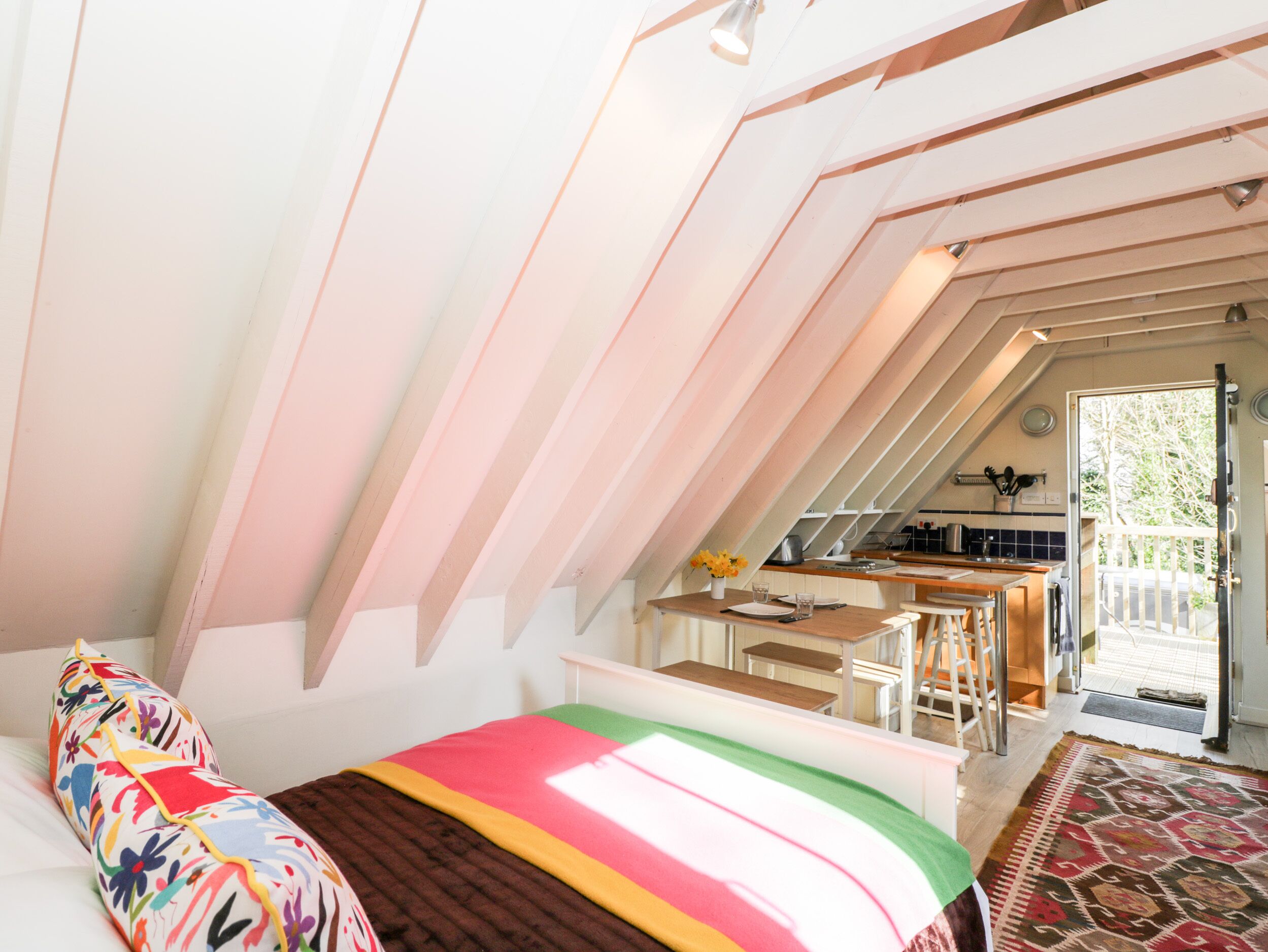 A bedroom with a sloped ceiling, a bed with a colorful floral pillow, a bedside table, and a view of a kitchen and balcony.
