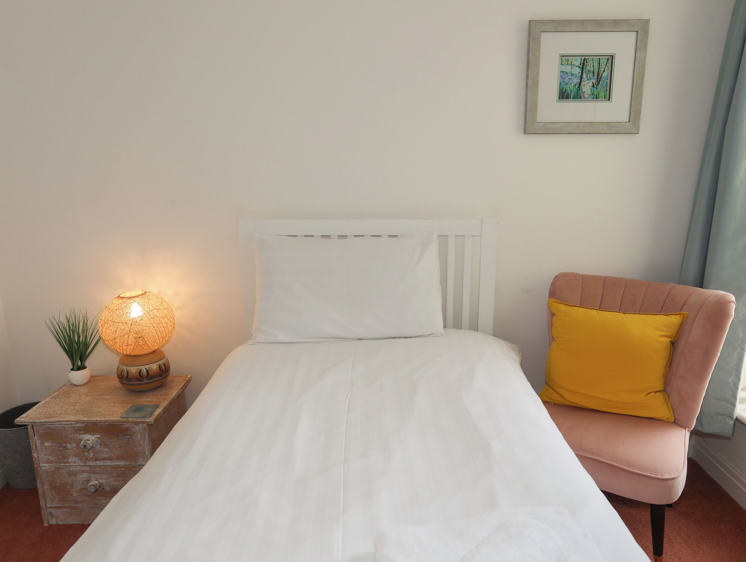 A bedroom with a bed, a bedside table with a lamp, a chair, and a framed picture on the wall.