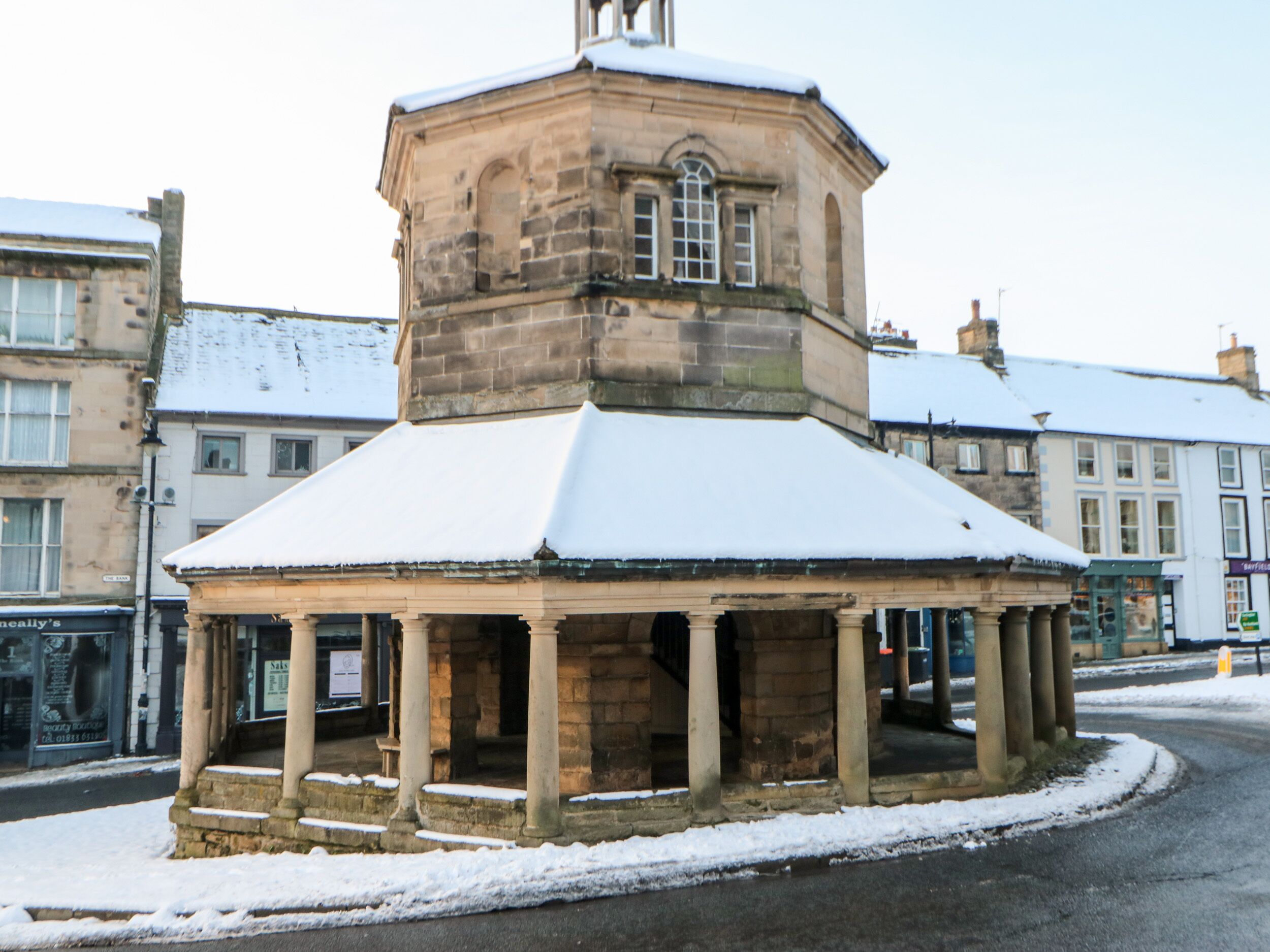 A stone gazebo with a snow-covered roof, surrounded by buildings.
