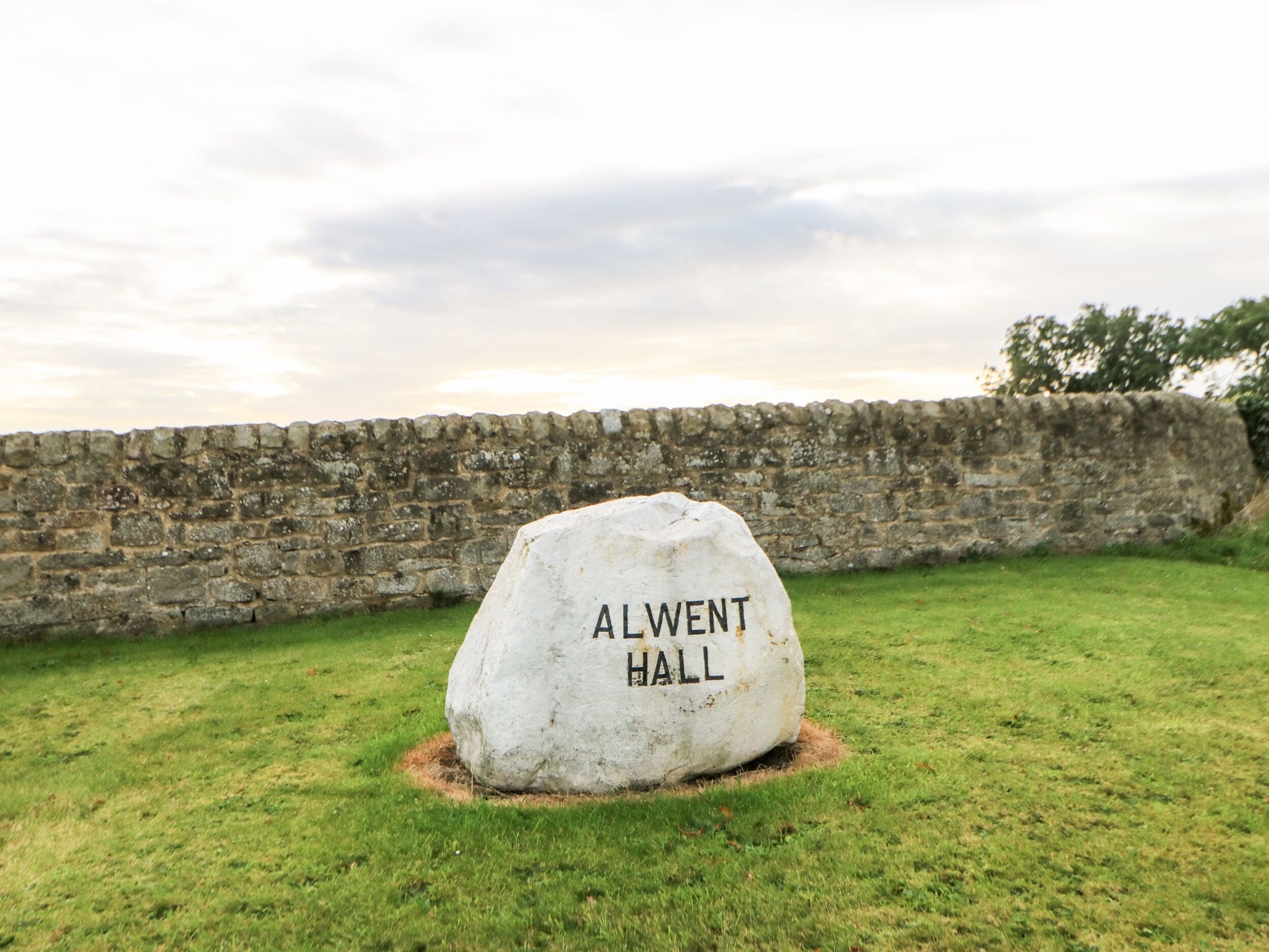 A large stone with 'ALWENT HALL' inscribed on it, surrounded by grass and a stone wall.
