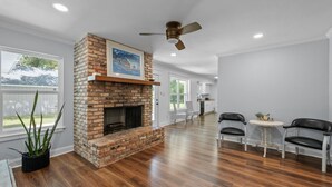 Un salón comedor amplio con pisos de madera, chimenea de ladrillo, ventilador de techo y una zona de comedor con dos sillas y una mesa pequeña.