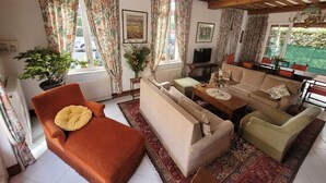 Un salon confortable avec un ensemble de canapés, une table basse et un téléviseur. On y trouve des rideaux à motifs floraux, une plante en pot et un radiateur.