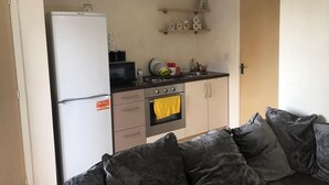 A compact kitchen with a refrigerator, microwave, stove, and a sink. There are cabinets and a countertop with various items. A couch with dark cushions is in the foreground. - Immaculate 1-bed Apartment in Stoke-on-trent (Stoke-on-Trent)
