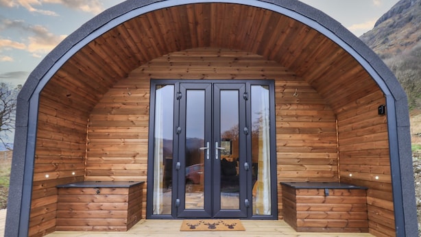A wooden cabin with a large arched entrance, a sliding glass door, and a small porch area with benches. - The Stag - Crossgate Luxury Glamping (Penrith)