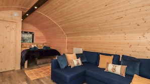 A cozy cabin interior with a wooden ceiling, a blue sectional sofa, and a bed with a forest-themed wall hanging. - The Stag - Crossgate Luxury Glamping (Penrith)