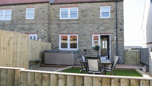 A two-story stone house with a tiled roof, a wooden fence, and a patio with a hot tub and outdoor seating.