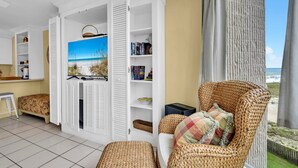 A room with a wicker chair, a bookshelf, a painting of a beach, and a view of the beach through an open door.