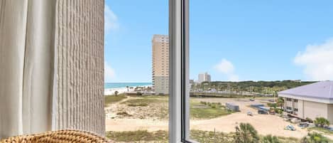 Une vue depuis une fenêtre donnant sur une plage, un parking et un bâtiment doté d’un toit plat.