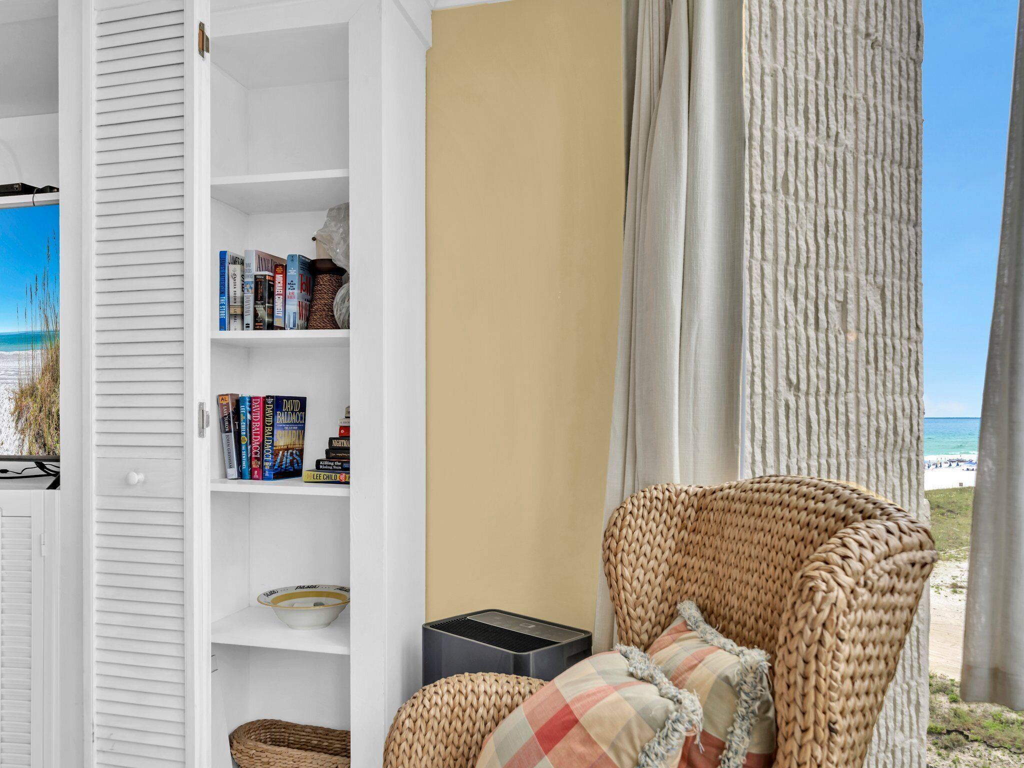 Ein Zimmer mit Bücherregal, einem Korbstuhl und Blick auf den Strand.