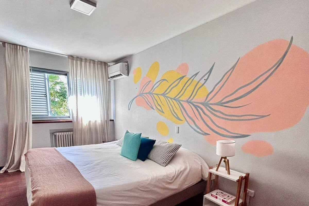 A bedroom with a bed, a window with curtains, a radiator, a wall with a leaf painting, and a bedside table with a lamp.
