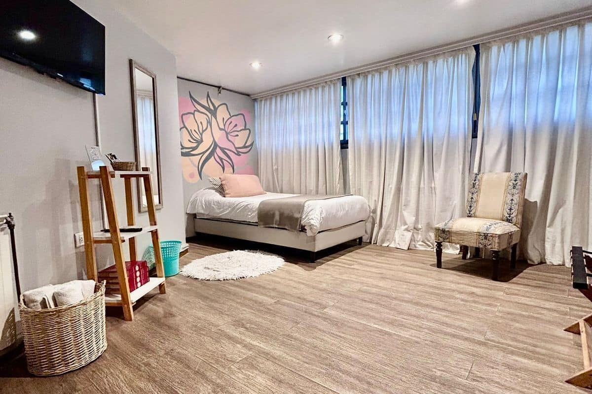 A bedroom with a bed, a chair, a TV, a wall-mounted shelf, and a large window with curtains.