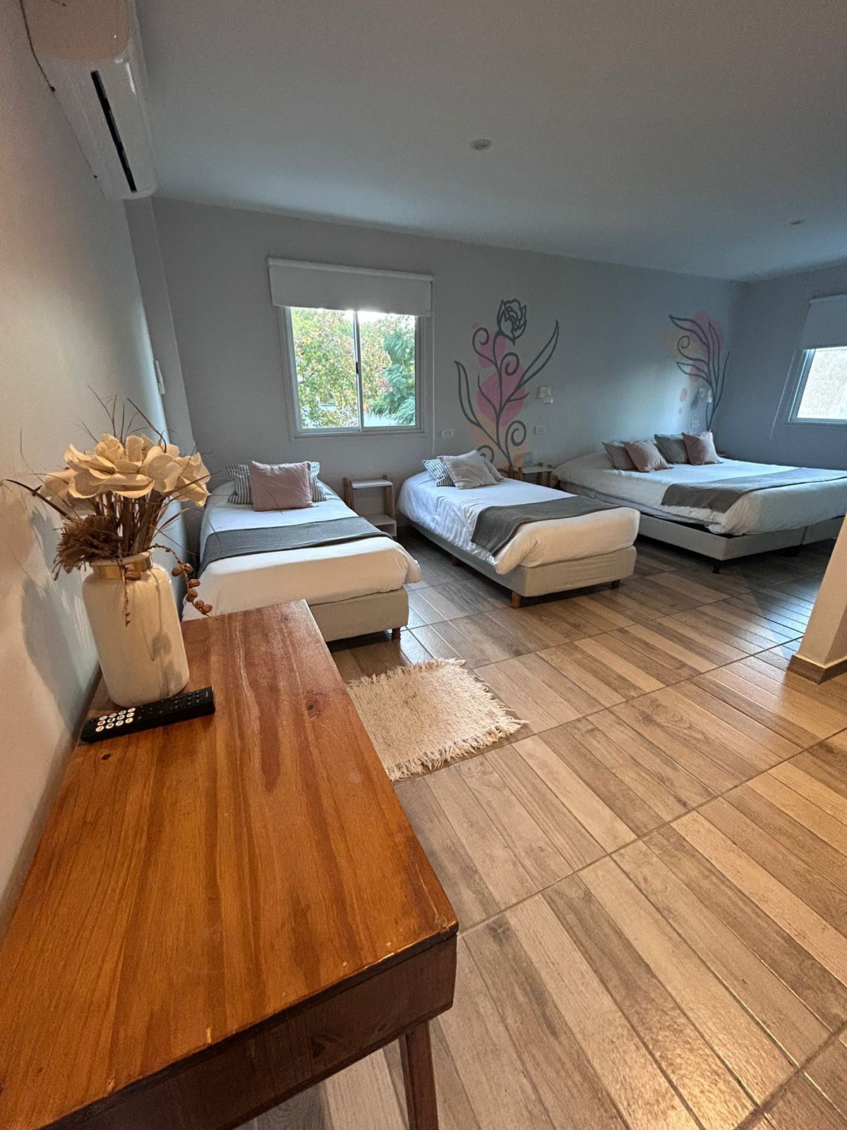 Quarto com duas camas, uma mesa de madeira, um vaso com flores secas e uma janela com vista para árvores.