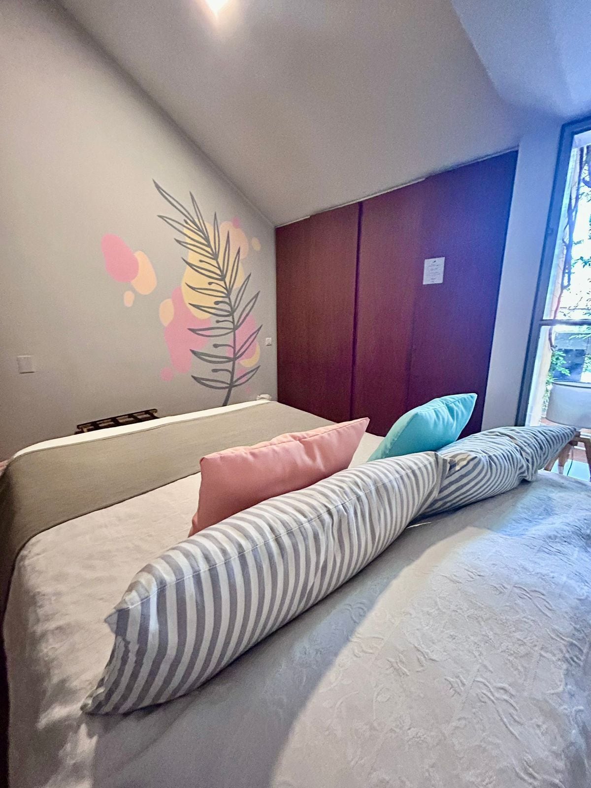 A bedroom with a bed, pillows, a wall with a leaf design, and a window.