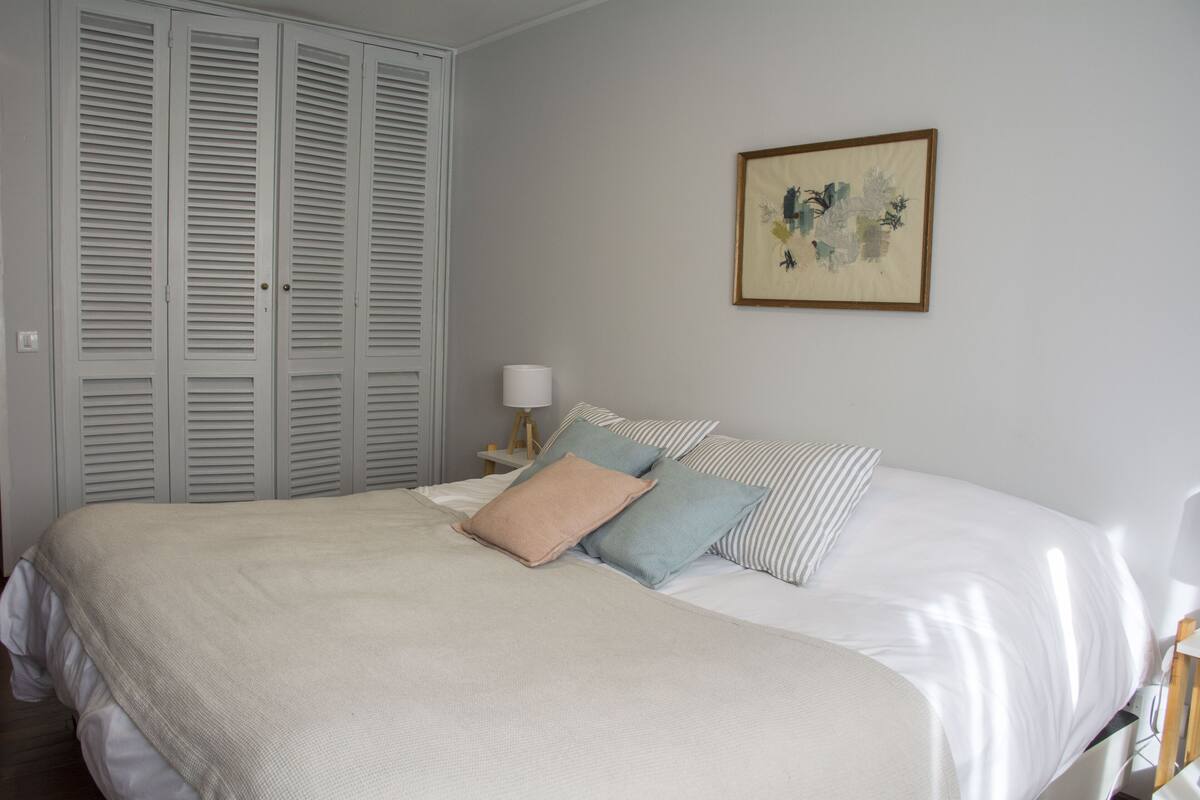 A neatly made bed with a white comforter and pillows, a bedside table with a lamp, and a large window with blinds.