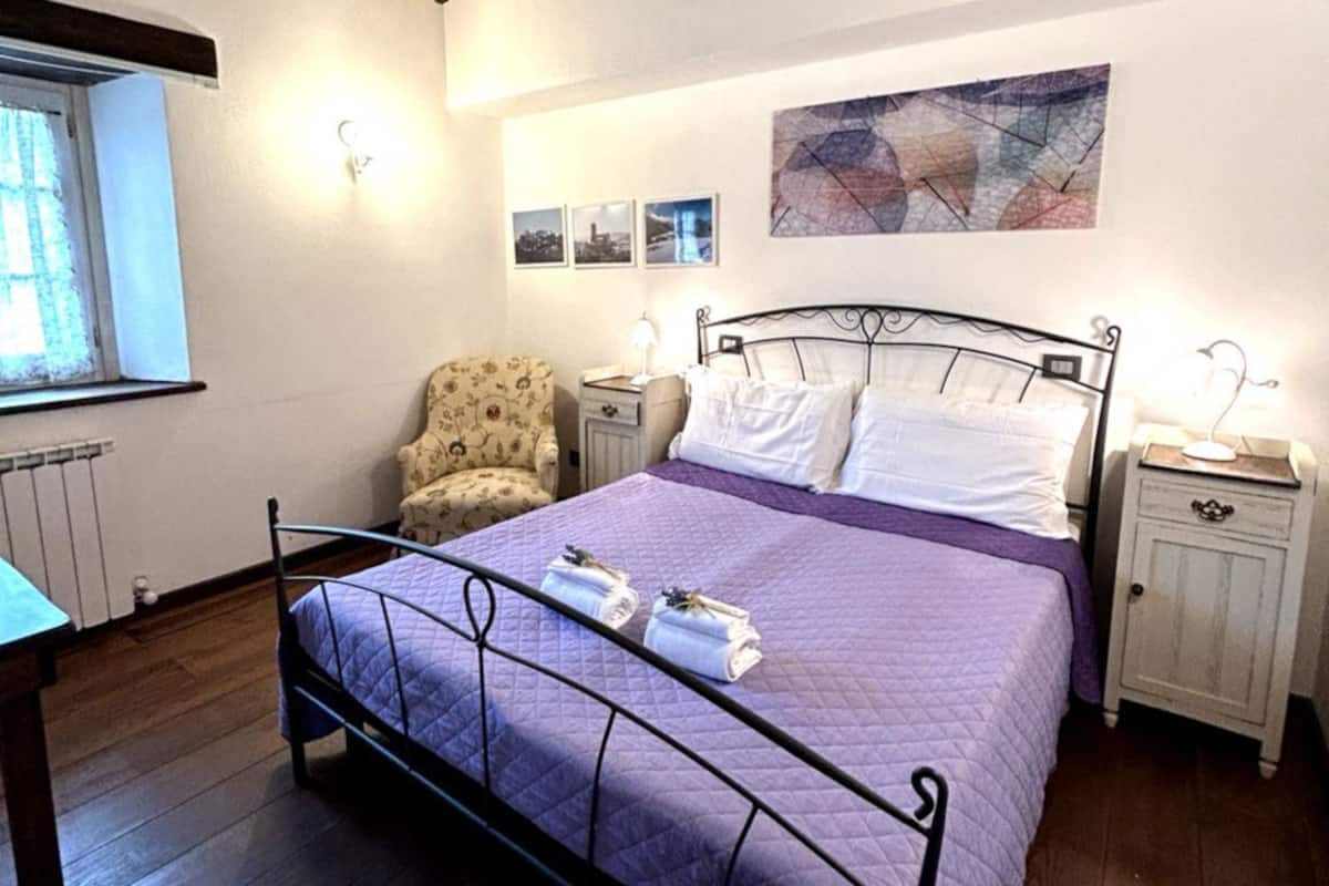 Un dormitorio con cama, una silla, una mesita de noche, una pared decorada con imágenes y una ventana.