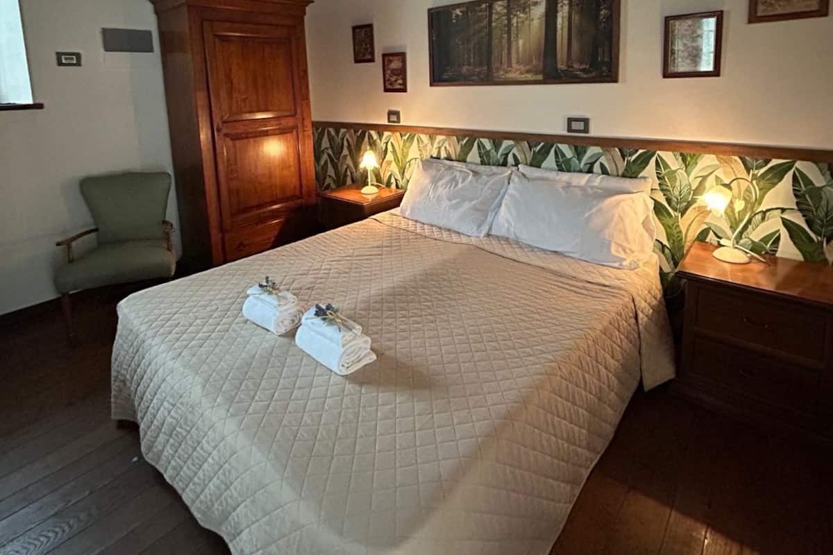 Un dormitorio con cama, cabecera de madera, mesitas de noche, una silla y un cuadro en la pared.