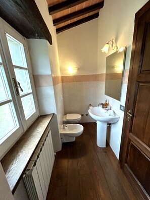 A bathroom with a toilet, bidet, and sink, wooden floor, and a window with blinds. - IL PIANETTO (Pieve Fosciana)