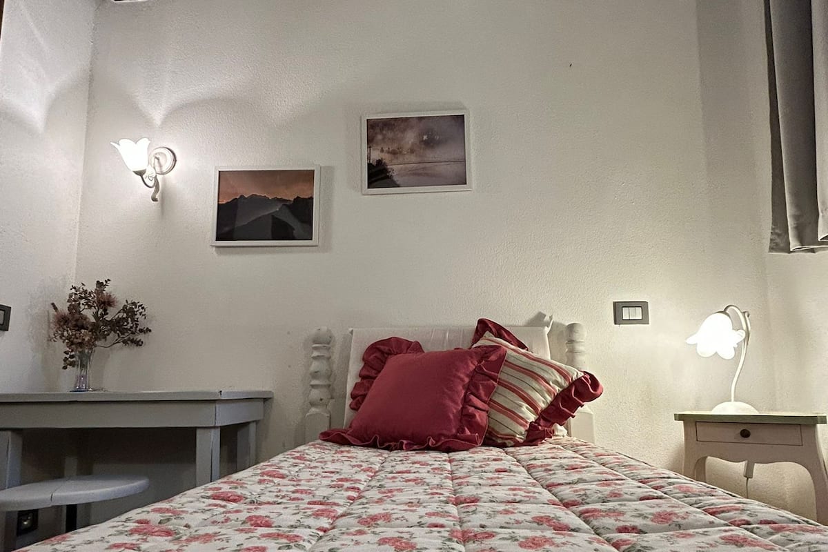 Un dormitorio con cubrecama floral, techo de madera, mesita de noche, lámpara y dos luces empotradas.
