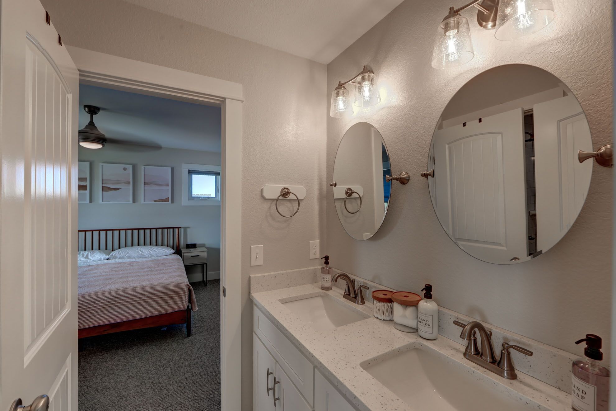 A bathroom with two sinks, a large mirror, and a toilet. There is a bed visible in the adjacent room.