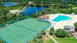 Ein Tennisplatz neben einem Schwimmbad mit überdachter Sitzgelegenheit, umgeben von Grünflächen und einem See.