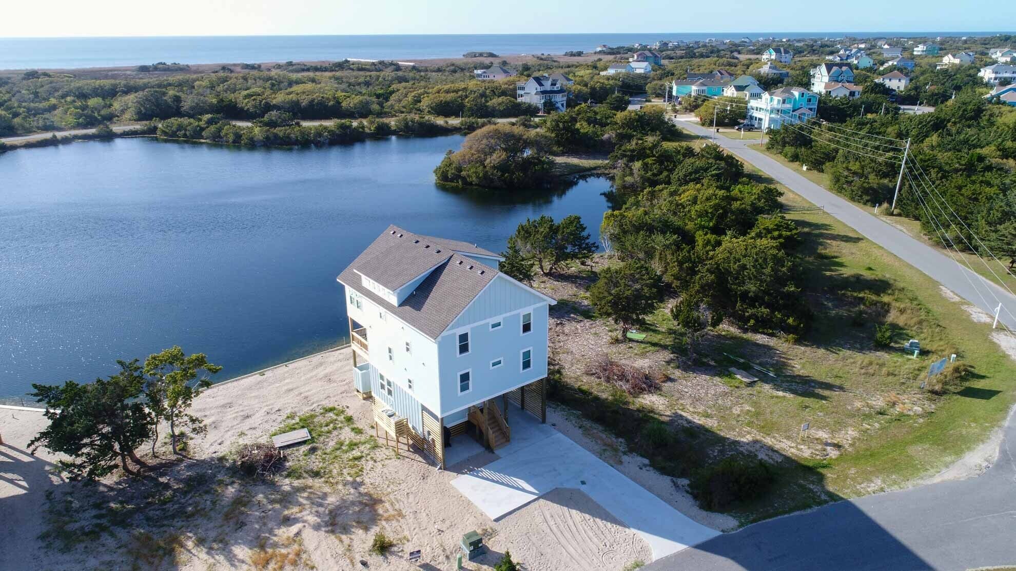 Een tweelaags huis met een groot erkerraam, gelegen aan een zandstrand met uitzicht op een rustig meer en een woonwijk in de verte.