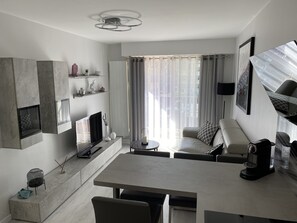 A modern living room with a flat-screen TV, a coffee table, and a sofa. There is a fireplace, a dining table, and a large window with curtains. - Beautiful new bright apartment facing south 100 m from the beach direct access (Cabourg)