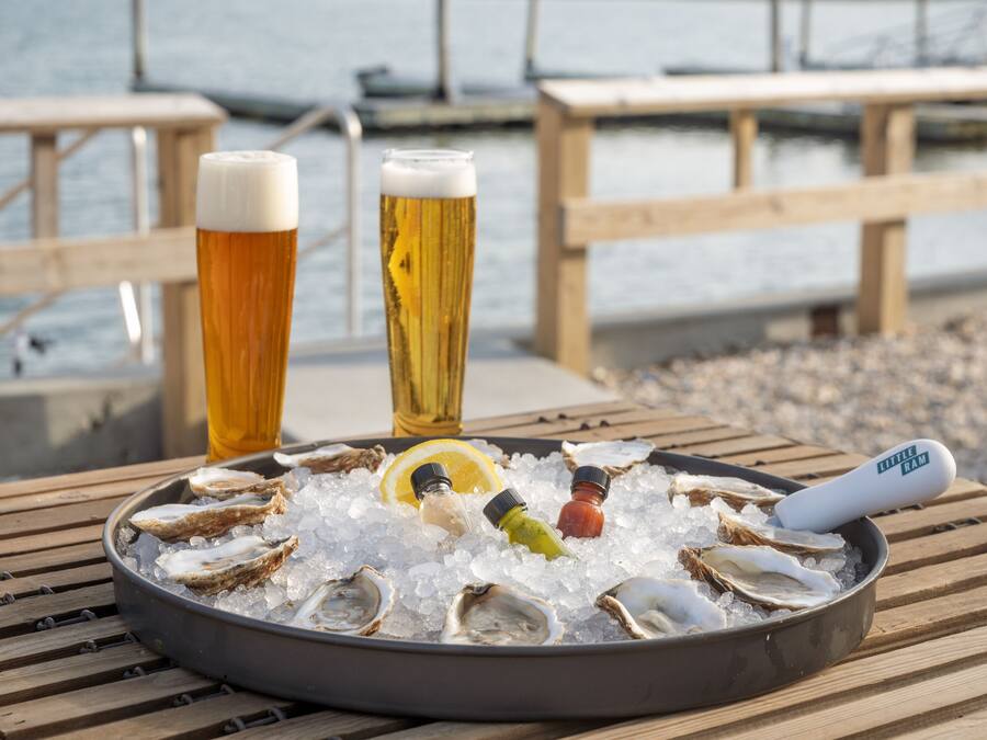 Una bandeja de ostras sobre hielo, con dos vasos de cerveza y una botella de salsa en una mesa de madera junto al agua.