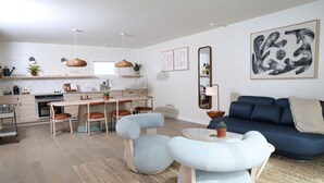 A modern living room with a blue sofa, light blue armchairs, a dining table, and a kitchen in the background. - The Shoals Suites and Slips (Southold)