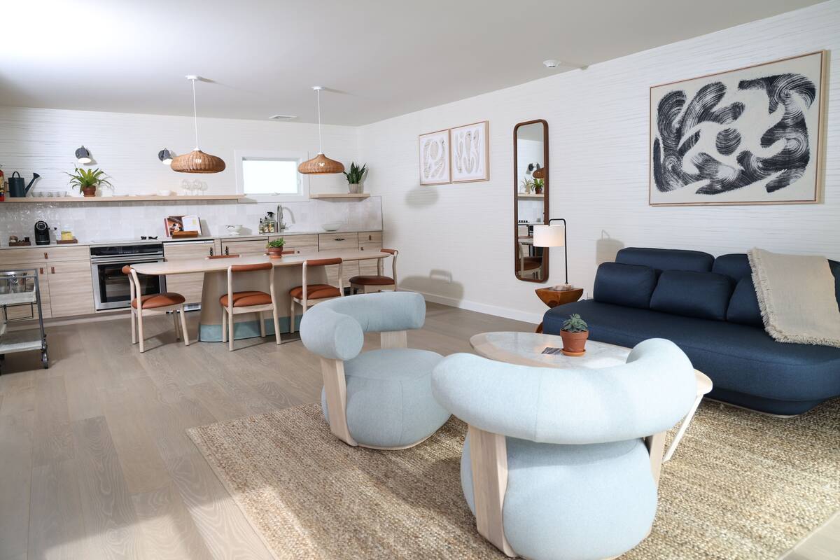 A modern living room with a blue sofa, light blue armchairs, a dining table, and a kitchen in the background.
