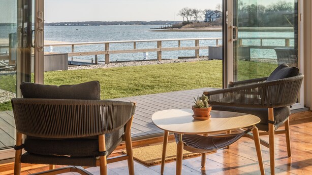 A modern outdoor seating area with two wicker chairs and a small table, overlooking a body of water and a wooden deck. - The Shoals Suites and Slips (Southold)