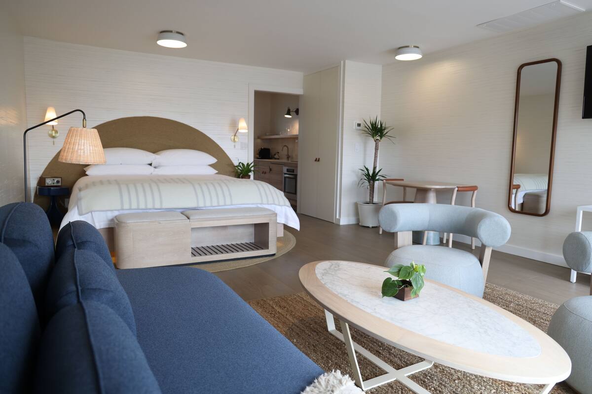 A modern hotel room with a large bed, a sofa, a dining table, and a kitchen area.