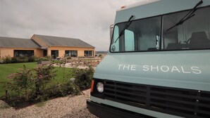 A light blue bus with "THE SHOALS" written on the front, parked in a driveway with a house and garden in the background. - The Shoals Suites and Slips (Southold)
